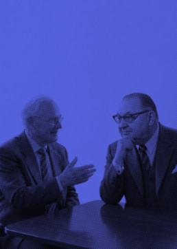 Jörg Koppenhöfer und Wilhelm Sander im Anzug im Gespräch an einem Tisch, künstlerisch in Blau dargestellt.
