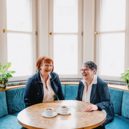Roswitha Engelmann und Thea Treffer lachen bei einem Kaffee in heller, freundlicher Atmosphäre