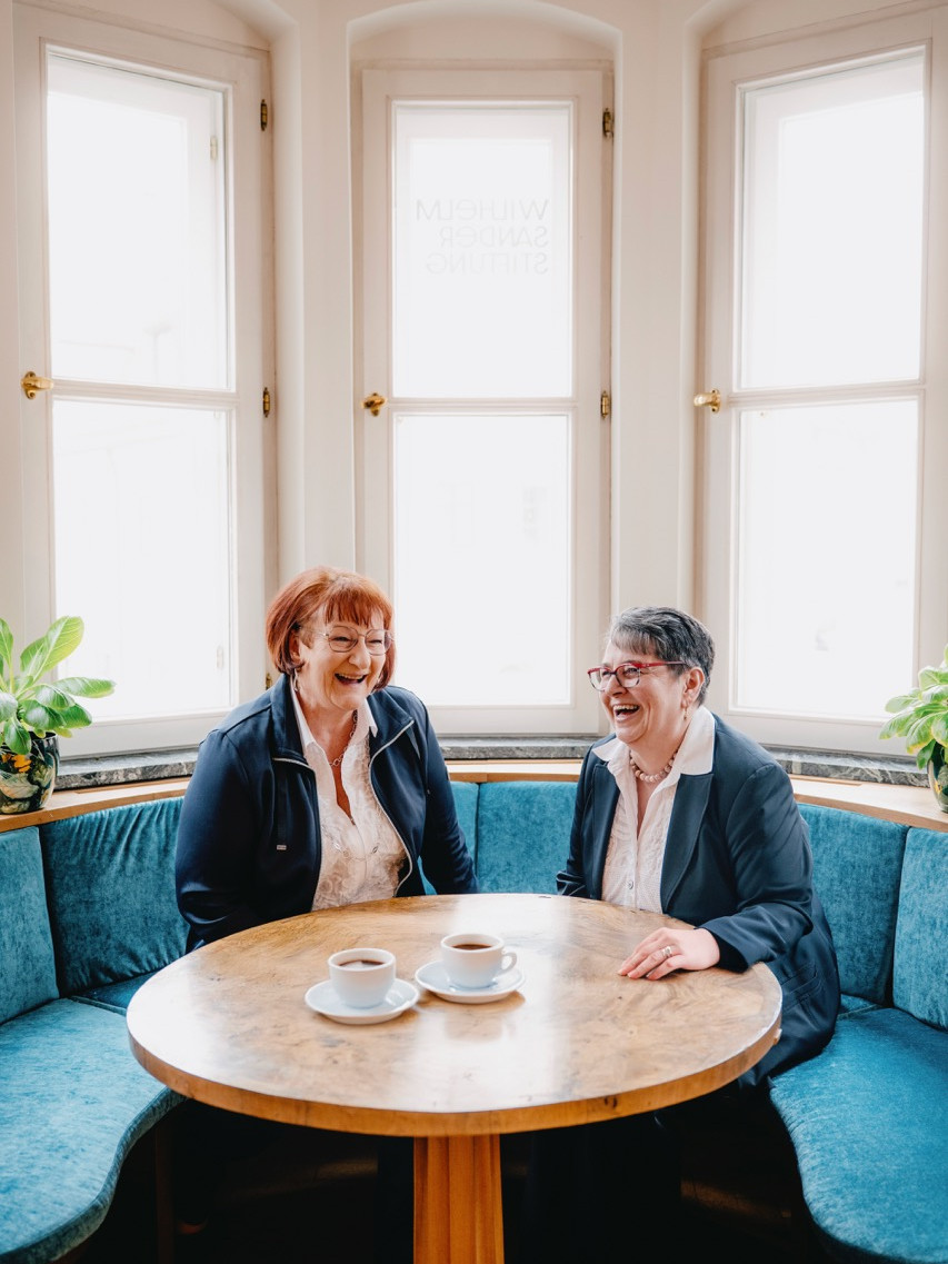 Roswitha Engelmann und Thea Treffer lachen bei einem Kaffee in heller, freundlicher Atmosphäre